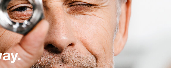 A photograph of a man with grey hair who looks through a screw-nut while inspecting it carefully.