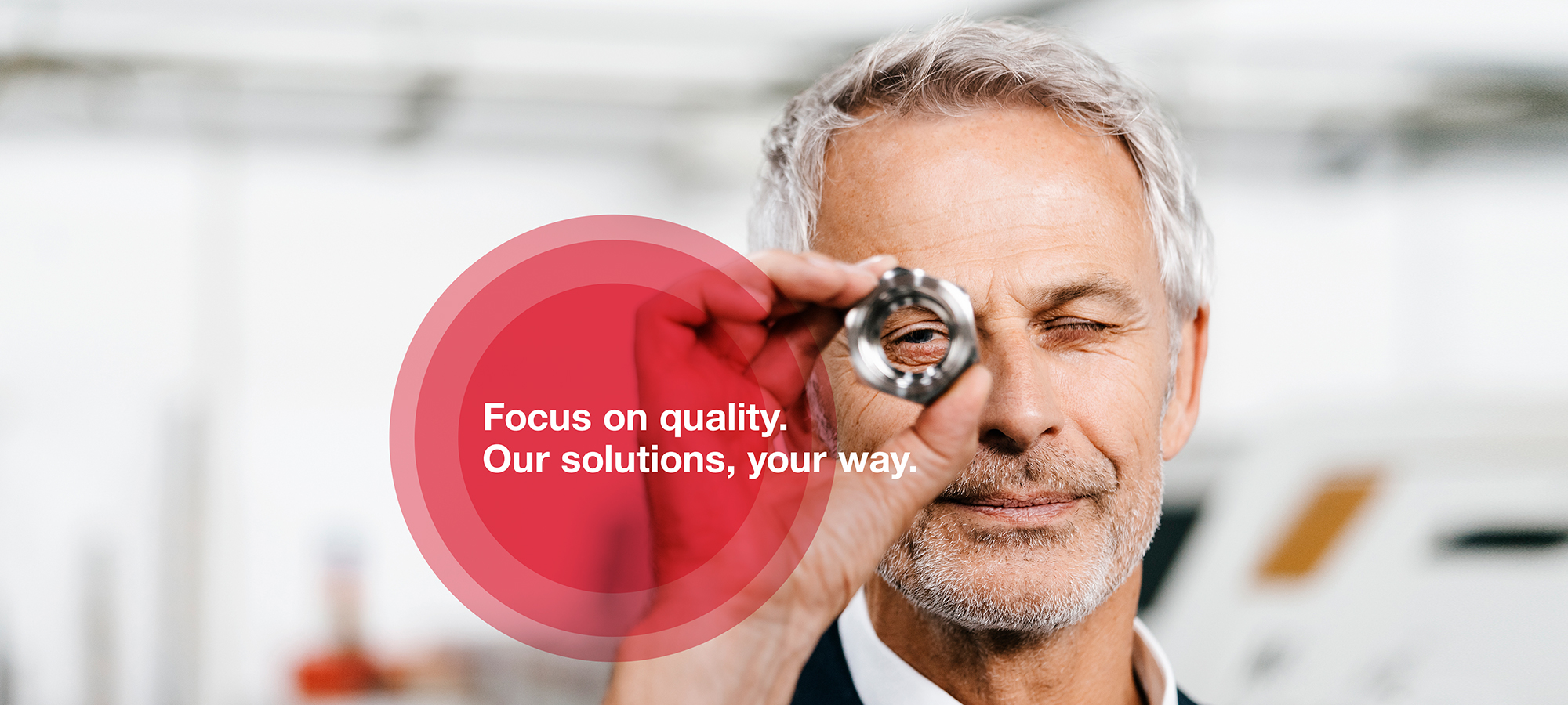 A photograph of a man with grey hair who looks through a screw-nut while inspecting it carefully.